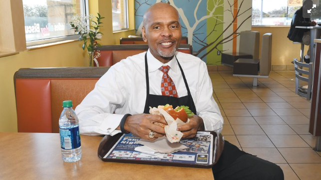 Roland Parrish, McDonald's franchisee, smiles while holding a burger at a table inside one of his McDonald's restaurants, wearing a black apron over a white shirt and tie. A photo of well-dressed man, Roland Parrish in a white dress shirt, red patterned tie, and black apron sits at a table inside a McDonald's restaurant, smiling warmly at the camera while holding a burger. A water bottle and a McDonald's serving tray with promotional materials rest on the table in front of him.