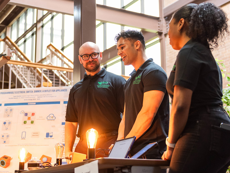 Photo of three UNT students stand in front of a table with illuminated industrial sized lightbulbs alongside a couple of ope laptop computers