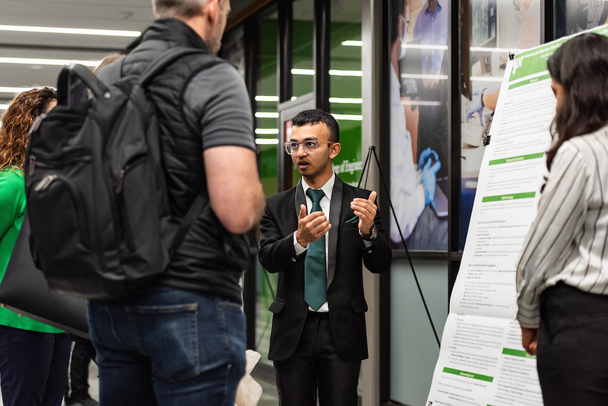 Photo of a student standing in front of a research poster explaining his work to others