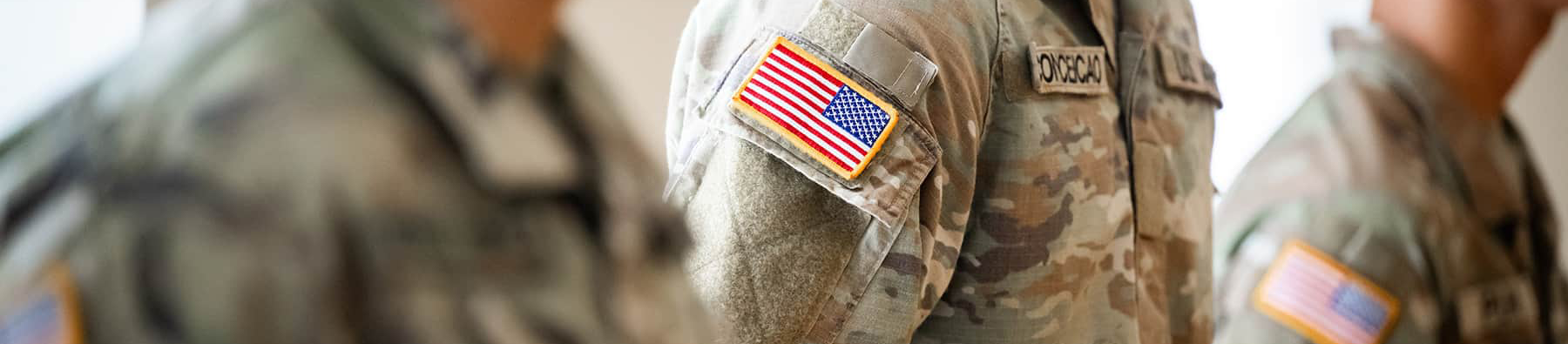 Top horizontal narrow cropped photo banner of three UNT DFW U.S. Army ROTC students in uniform standing alert in formation. The photo's selective focus is on the shoulder patch of the US flag.