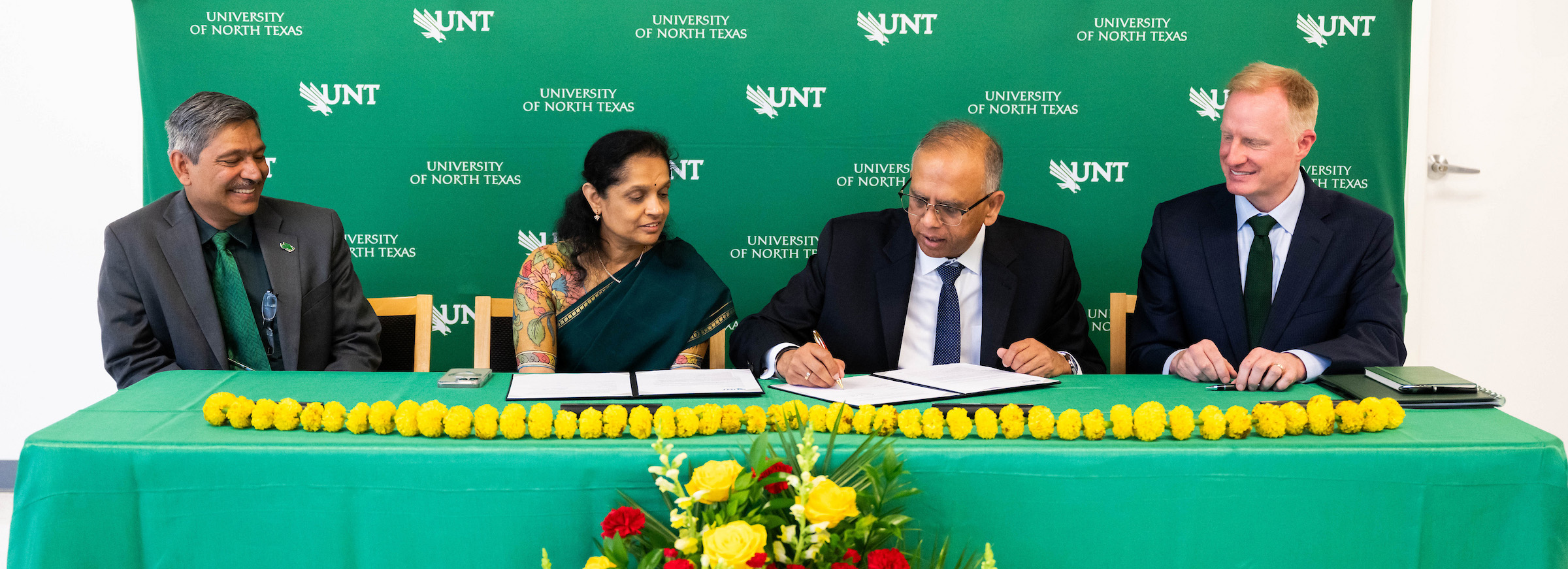 Top horizontal photo banner (from left to right) UNT College of Information's Dean Kinshuk, Anuradha Sinha with Vikas Sinha signing a sheet of paper next to UNT President Harrison Keller. All are sitting down at al long table. 