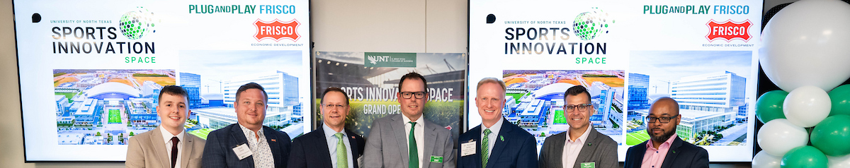 A group of seven men in business attire pose for a photo at the University of North Texas Sports Innovation Space event. Two large digital presentation screens and balloons in the background. Top horizontal narrow cropped photo banner of a group of seven men in business attire pose for a photo at the University of North Texas Sports Innovation Space event. Two large digital presentation screens and balloons in the background.