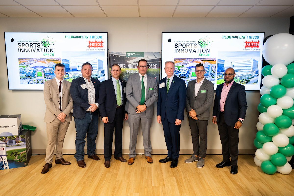 A group of men pose for a photo at UNT Sports Innovation Space event in Frisco.  Full photo of seven men stand together in front of screens that say Sports Innovation with the Frisco logo in the corner