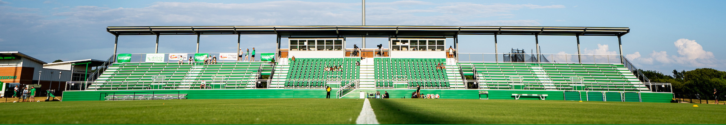 Top horizontal, narrow-cropped photo banner of a wide view of a UNT's green soccer stadium with mostly empty bleachers under a blue sky.  