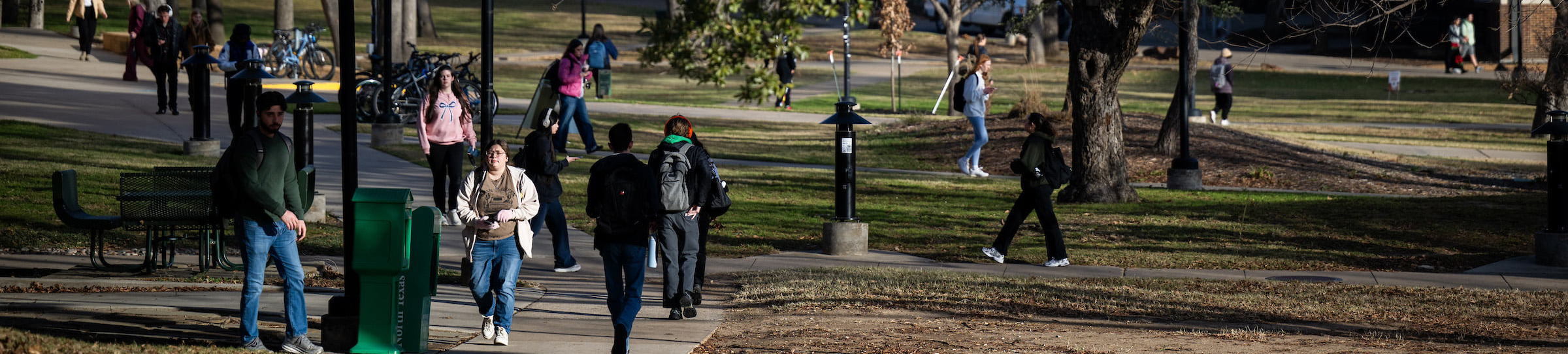 UNT students walking across a tree-filled campus with pathways and benches on a sunny day.\ Top, wide banner photo of UNT students walking across a tree-filled campus with pathways and benches on a sunny day.