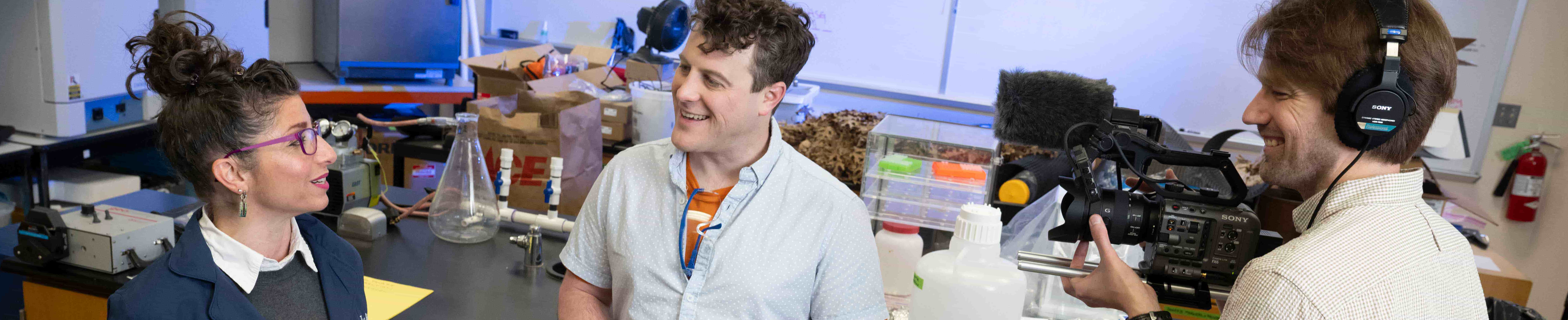 Top wide-narrow cropped banner photo of three people in a UNT College of Science laboratory: A lady and a man are talking while a third man films them with a camera.