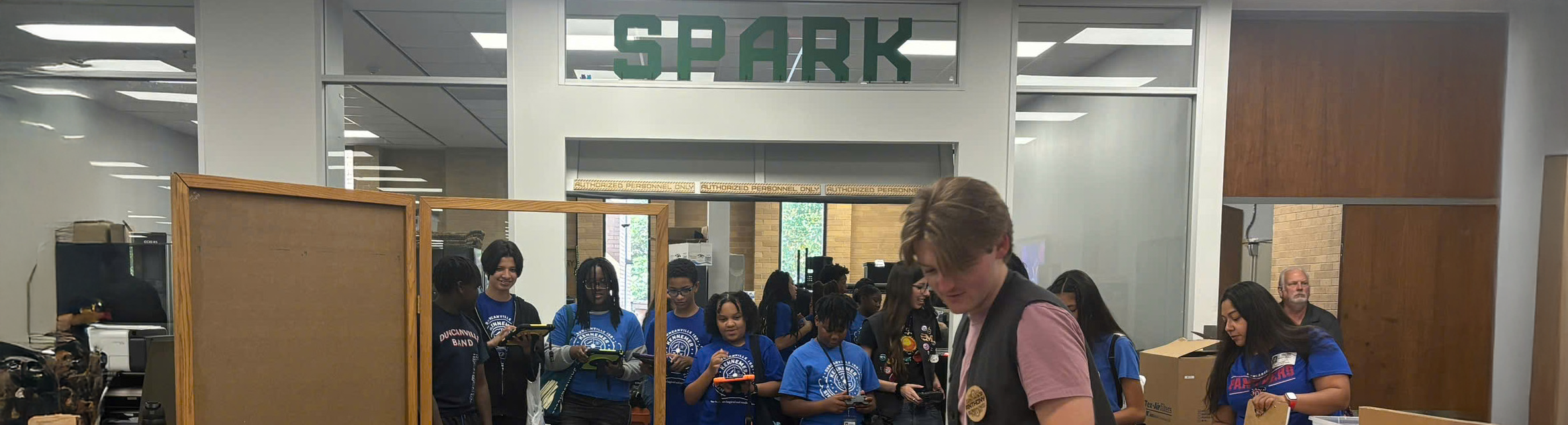 Photo banner of UNT's Willis Library’s Spark Makerspace. Showing visiting middle school students in blue shirts explore hand held controllers and tablets around activities while instructors assist in the background. Top horizontal, narrow-cropped photo banner of UNT's Willis Library’s Spark Makerspace. Showing visiting middle school students in blue shirts explore hand held controllers and tablets around activities while instructors assist in the background.
