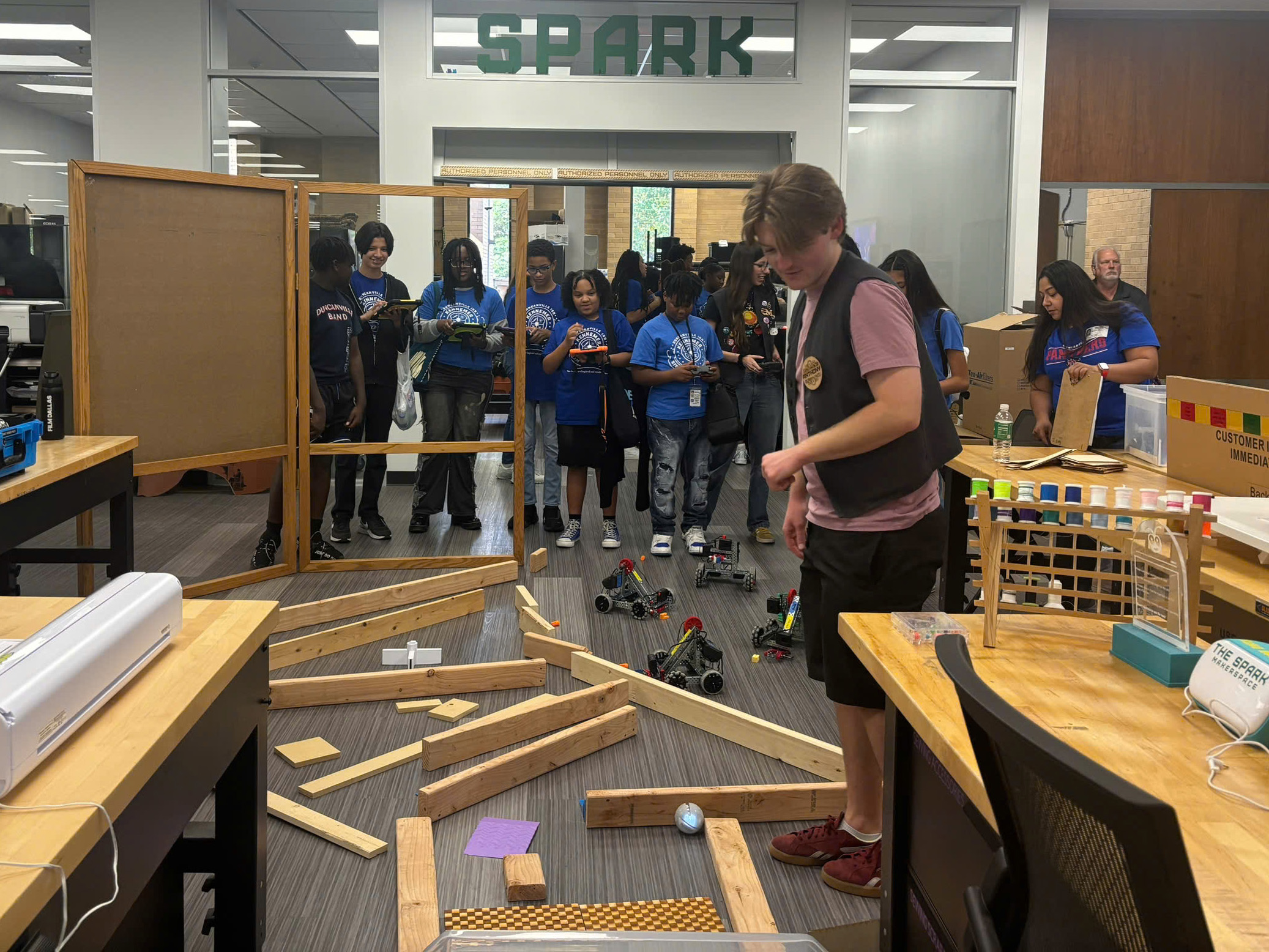 A group of young 7th grade students in blue stand in the background. They hold control panels for machines going through a makeshift obstacle course on the floor in front of them. A UNT library worker stands in the foreground watching the machines. Full photo of a group of young 7th grade students in blue stand in the background. They hold control panels for machines going through a makeshift obstacle course on the floor in front of them. A UNT library worker stands in the foreground watching the machines.