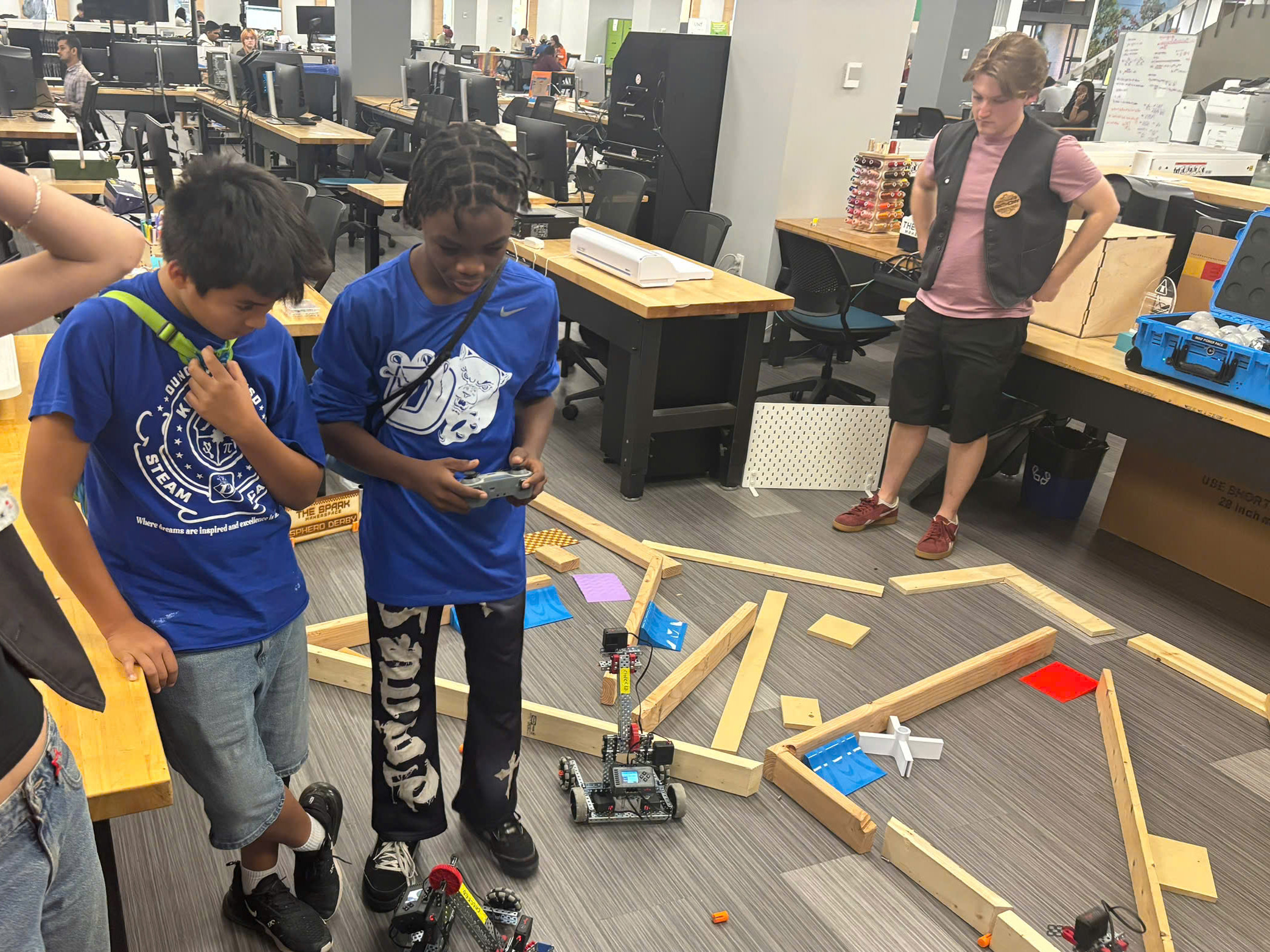 Two visiting middle school students in blue shirts control robots on the floor surrounded by a wooden barriers while a UNT staff member observes. Photo of two visiting middle school students in blue shirts control robots on the floor surrounded by a wooden barriers while a UNT staff member observes.