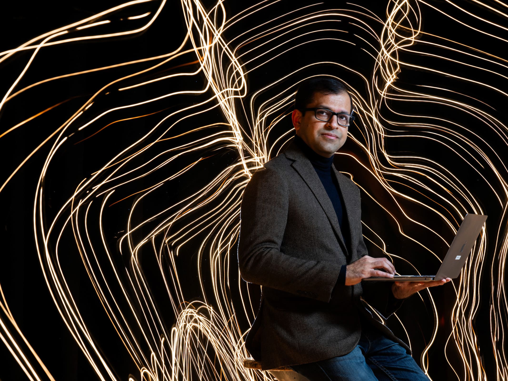 Photo portrait of UNT's Supreeth Shastri using a laptop with dynamic penlight trails repeating his outline shape around him frozen in time in the background.