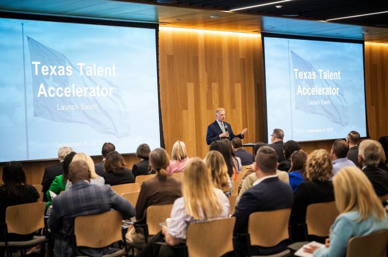 Photo of UNT President Harrison Keller holding a microphone speaking to an audience. On either side are large screens with a picture of the Texas State Flag and the words Texas Talent Accelerator Launch Event.