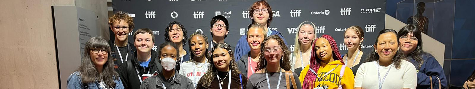Top horizontal narrow cropped photo banner of a group UNT students who attend the 2025 Toronto International Film Festival standing indoors in front of a large black step and repeat backdrop showing the festival's corporate sponsors logos in white.