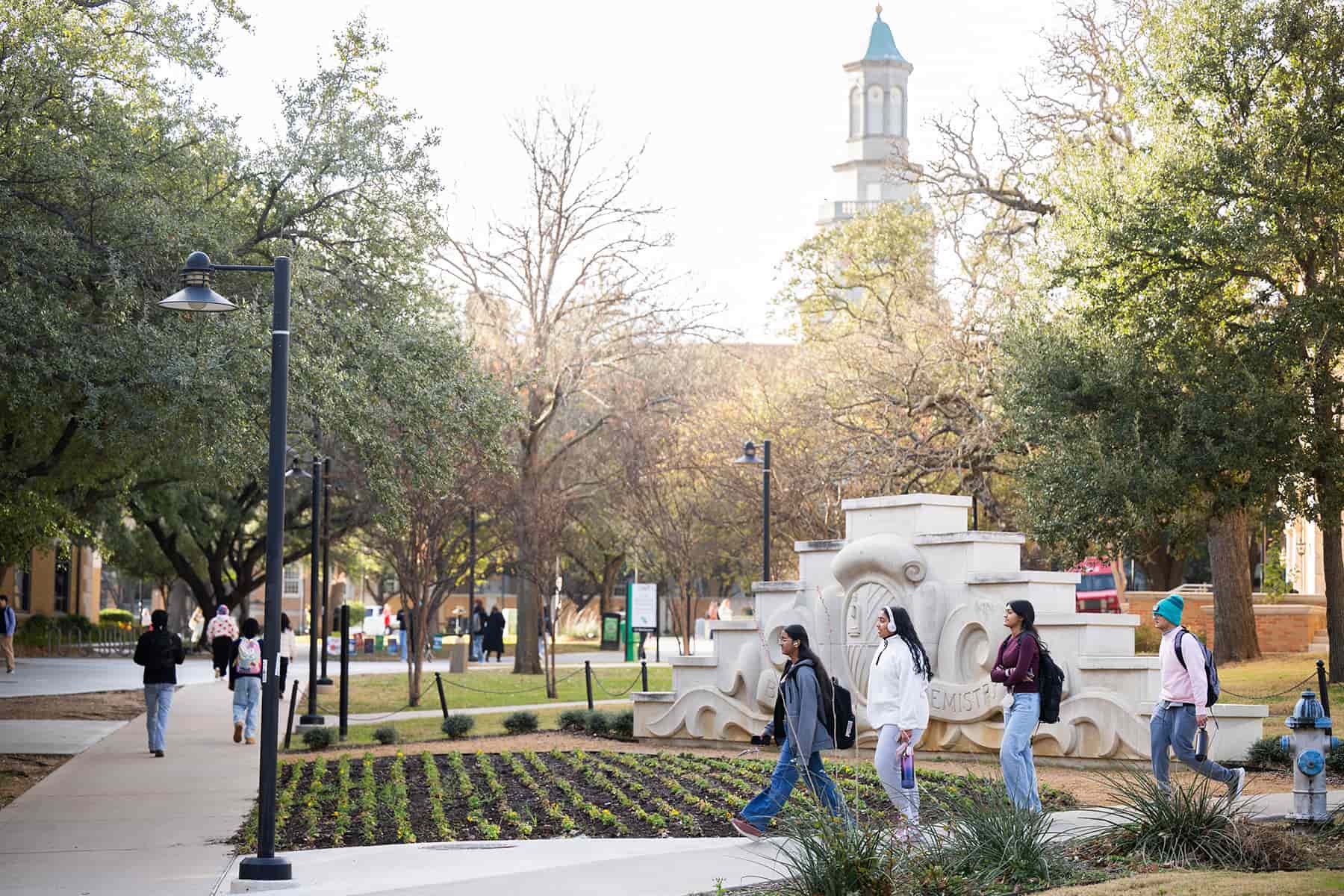 Full photo showing students walking across the UNT Denton campus near a decorative stone monument, with tree-lined pathways and the iconic UNT clock tower visible in the background.
