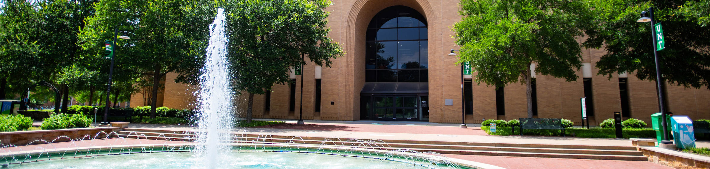 Top, wide banner photo of Willis Library fountain at the University of North Texas (UNT) campus, Denton, TX next to the Willis Library when there is a row of trees against the walls between the main entrance.