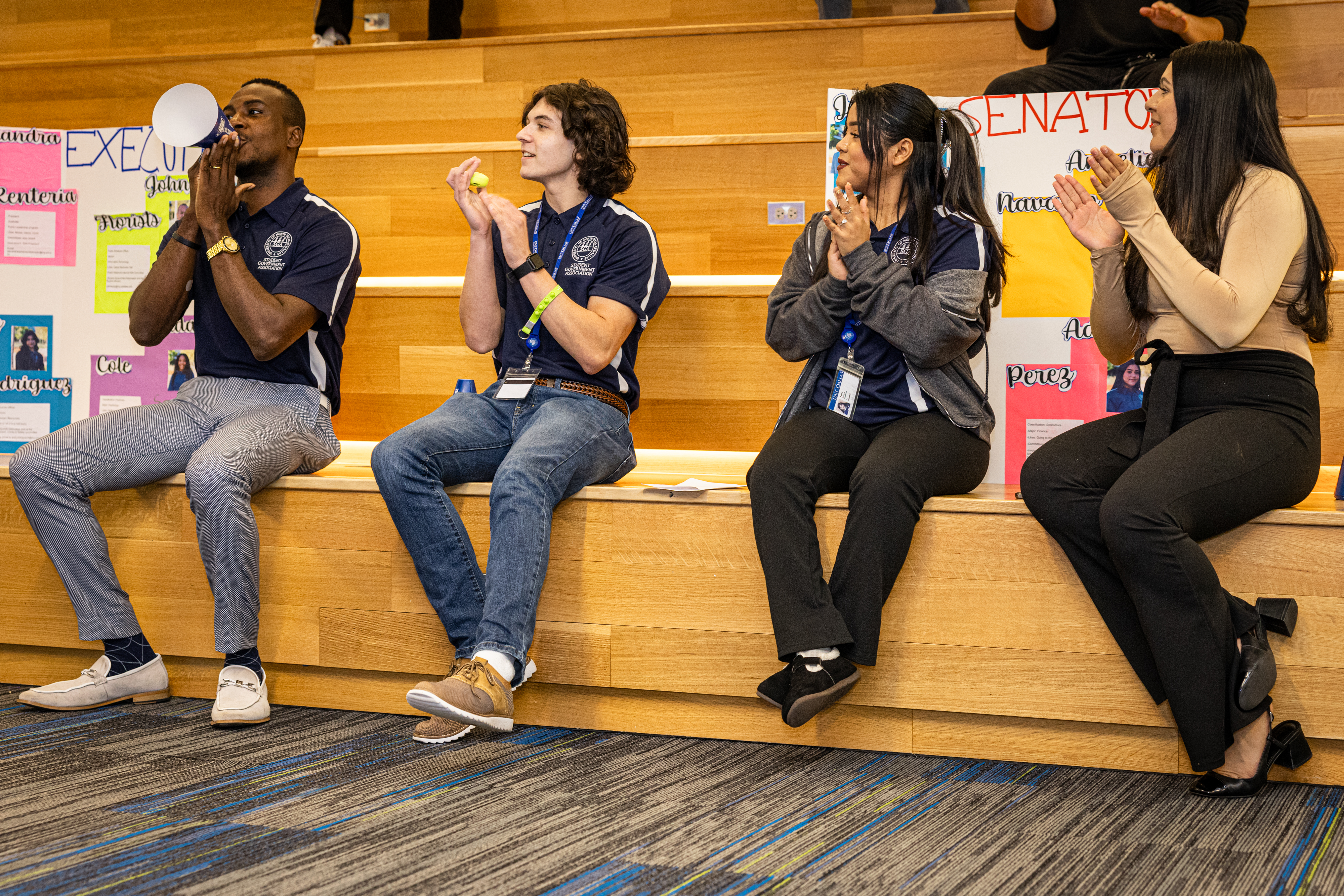 A Photo showing 4 UNT Dallas Student Government Association members seated on a wooden styled bleacher arrangement introducing themselves to fellow students