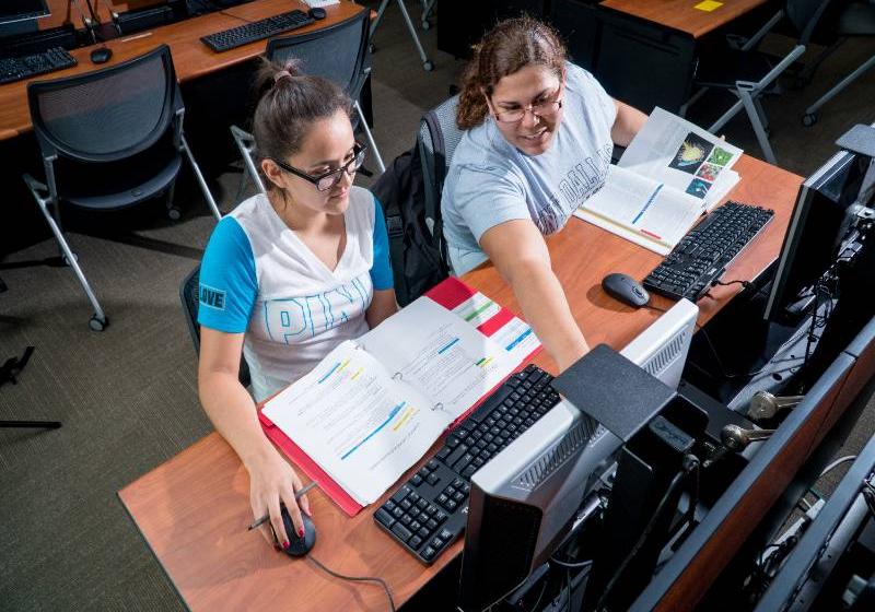 UNT Dallas students working together at their own in a computer lab. A photo of two more UNT Dallas students working together at their own computers with open textbooks in a computer lab.