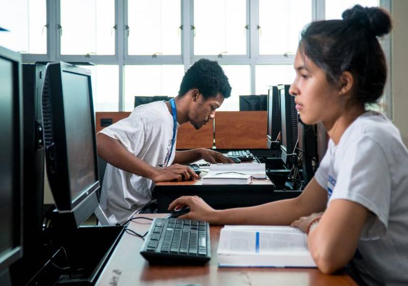 Students using computers A photo of two UNT Dallas students working on computers and reading books in a computer lab.