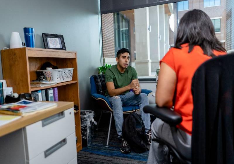 A photo of a young man sits and talks with a female counselor in an office with a large window.