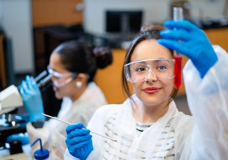 A photo close-up of one UNT Dallas female student wearing safety goggles, a translucent coat over her normal wardrobe with medical safety gloves holding a test-tube with a red liquid in one hand and in her other a long stem cotton swab. In the immediate background, but out of focus, is an other student examining something through a microscope dressed in the same way.