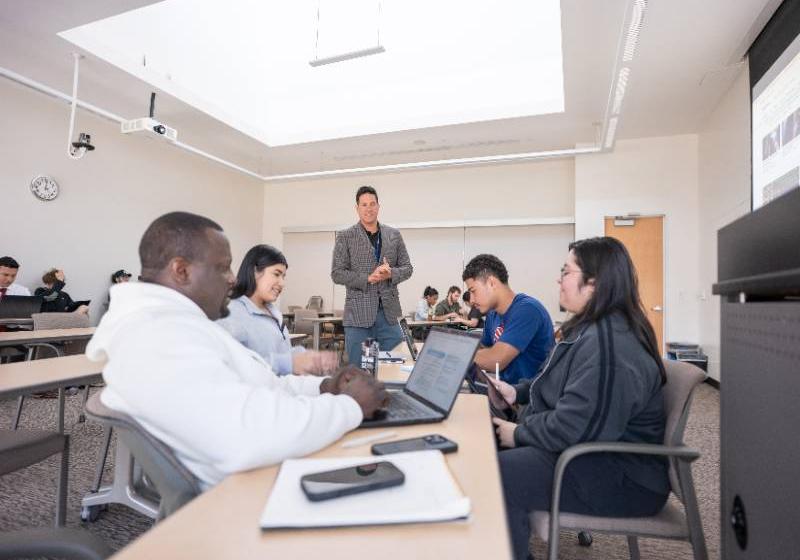 A photo of seated UNT Dallas business students with their open laptops on long tables engage with their professor standing during an interactive class activity.