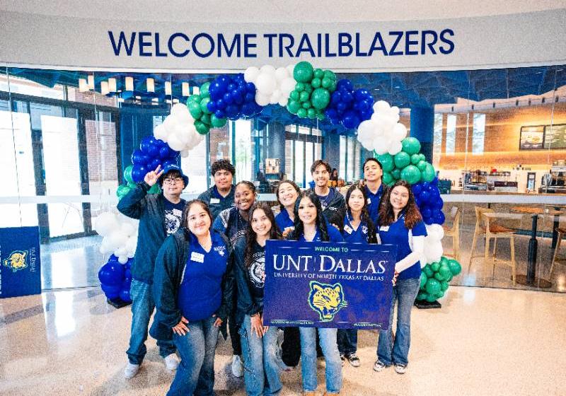 A Photo of 11 UNT Dallas students in one group with one on the girls in the front right is hold up a very large card with welcome to UNT Dallas printed on it.