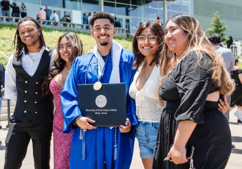 Photo with a graduate wearing his cap and gown together posing with 4 friends celebrate their achievements and experiences outside the Texas Trust CU Theater 