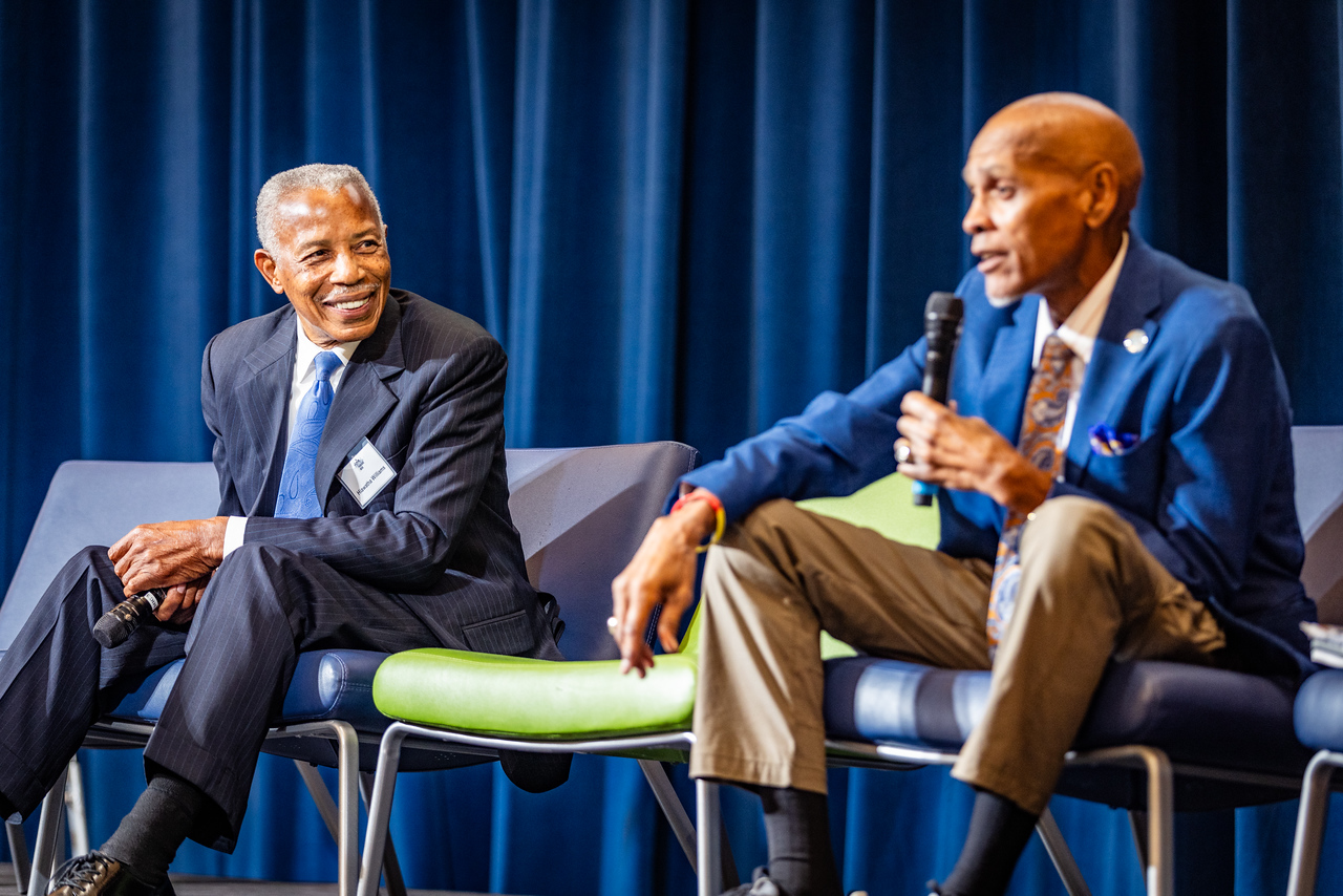 Photo of Hiawatha Williams (left), founder of the Williams Chicken Restaurant Chain (not holding a microphone), both sitting down in chairs infront of a blue curtain, speaks at a UNT Dallas School of Business Franchise Week Event