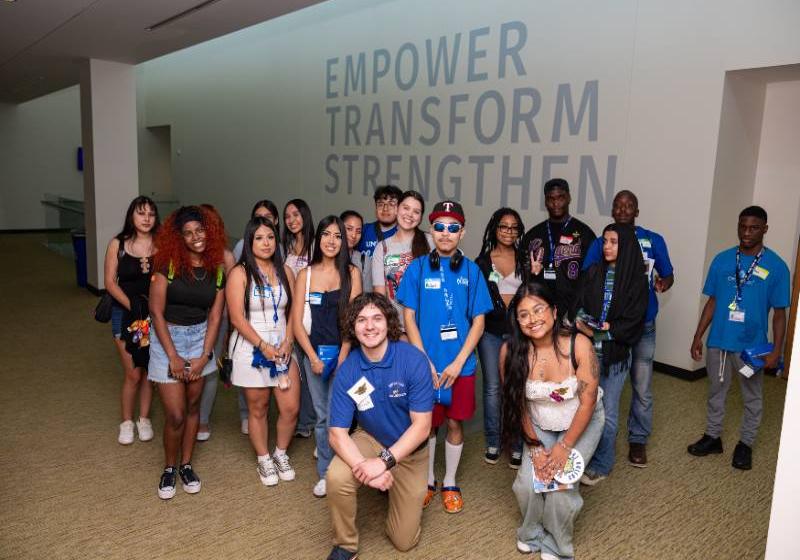 A large group of UNT Dallas students posing inside in front of a wall that has the largely visible words: EMPORE. TRANSFORM. STRENGTHEN. painted on it.