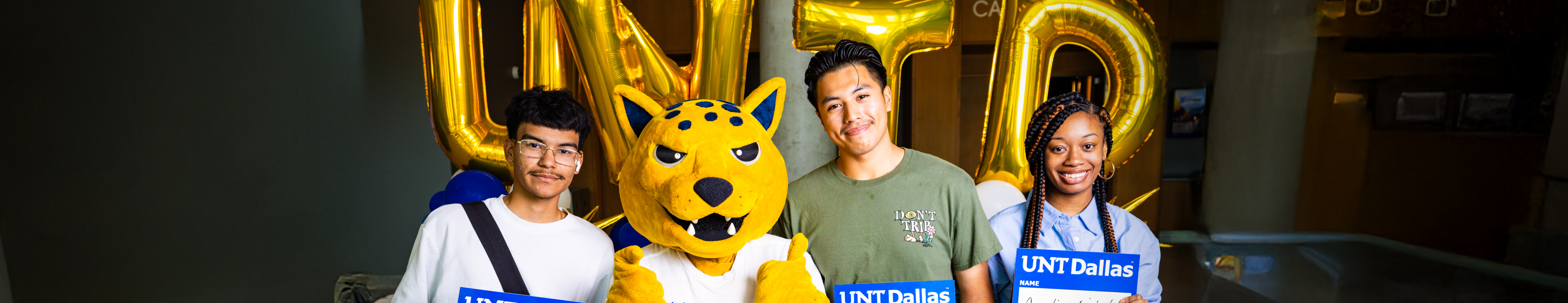 Top horizontal cropped photo banner of 3 new UNT Dallas students on their first day of school posing with the jaguar mascot