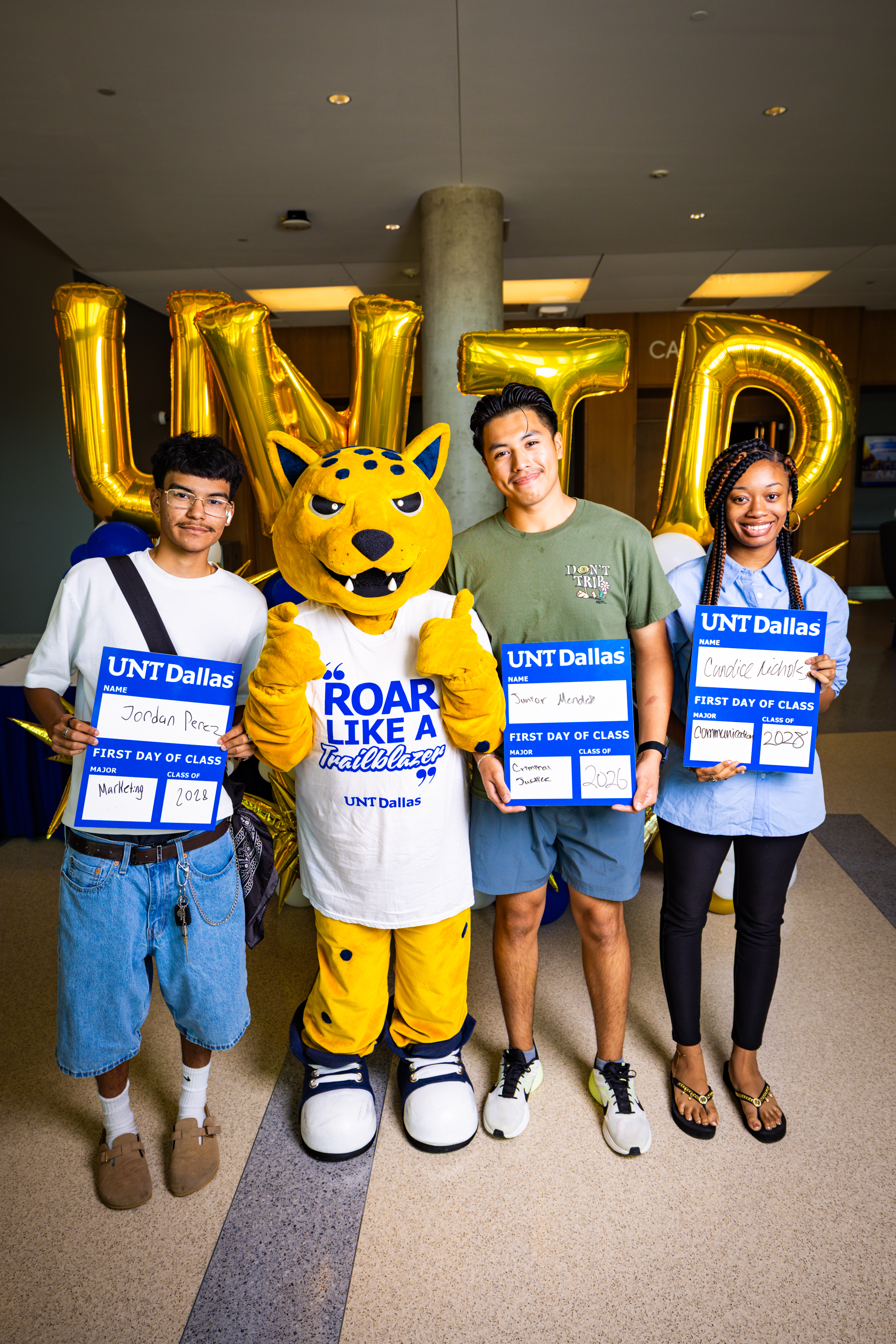 3 new UNT Dallas students on their first day of school posing with the jaguar mascot standing in the middle. The 2 males and 1 female freshman are holding an erasable laminated card with their name, major and class year. Behind them are 4 golden letter shaped balloons spelling out UNTD