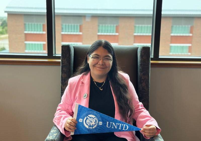 UNT Dallas's Julisa Morones Luna Photo of a woman in a pink blazer as she sits in a chair holding a blue UNT Dallas pennant, smiling in front of a window.