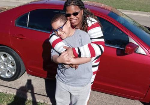 Photo of Yolanda Lewis with her arms wrapped around her grandson, Antonio. They are both standing outside in front of a red car 