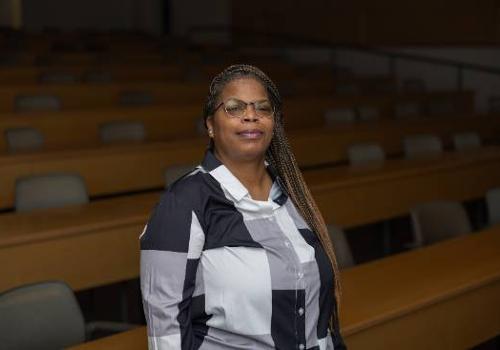 Photo of Yolanda Lewis at UNT Dallas one week before commencement standing by herself in a dark and empty lecture room in front of rows of connected desks with chairs