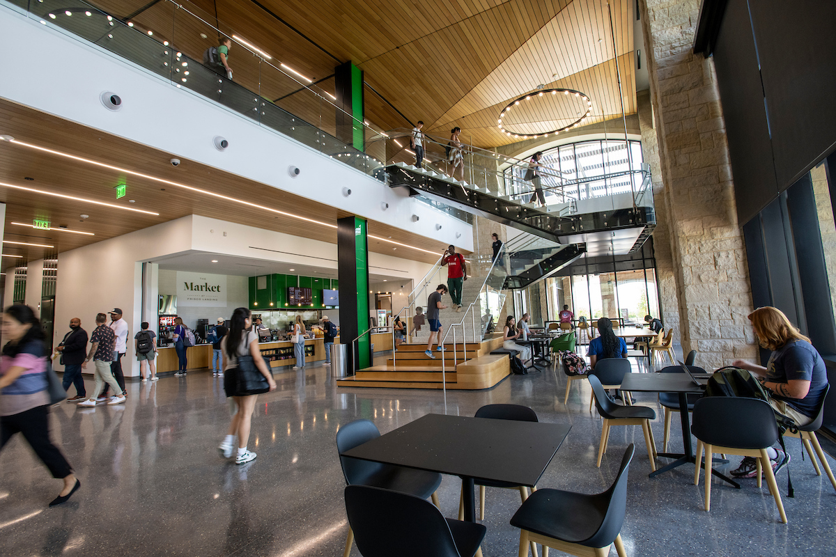 Students walks through the UNT at Frisco lobby area, sit at tables working, and walk on the central staircase. A photo of the UNT at Frisco lobby. Students walks through the area, sit at tables working, and walk on the central staircase. There is a central green pillar and a market open to the left.
