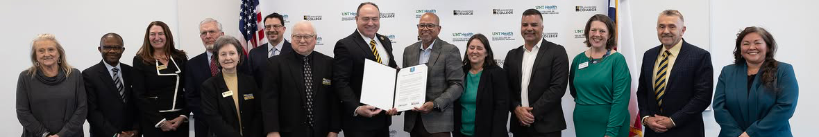 Photo of fourteen people standing behind their official signing ceremony table, representing both Weatherford College (on the left) and UNT Health Texas College of Osteopathic Medicine (on the right), celebrates their signed agreement. Top vertical photo banner of a cropped long horizontal photo of fourteen people standing behind their official signing ceremony table, representing both Weatherford College (on the left) and UNT Health Texas College of Osteopathic Medicine (on the right), celebrates their signed agreement. In the middle, both presidents, Dr. Tod Allen Farmer from Weatherford College and Dr. Kirk Calhoun from UNT Health, are holding up the document in an open black folder together. Behind them, on a wall, is a banner displaying both schools' repeated logos in a stair-stepped fashion in the background.
