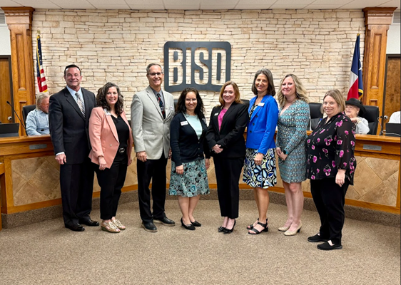 Photo of nine professionals — a mix of men and women in business attire — pose together smiling in an official BISD (Birdville Independent School District) boardroom. They stand in front of a stone accent wall displaying the BISD logo. An American flag and a Texas state flag are visible in the background, along with boardroom seating.