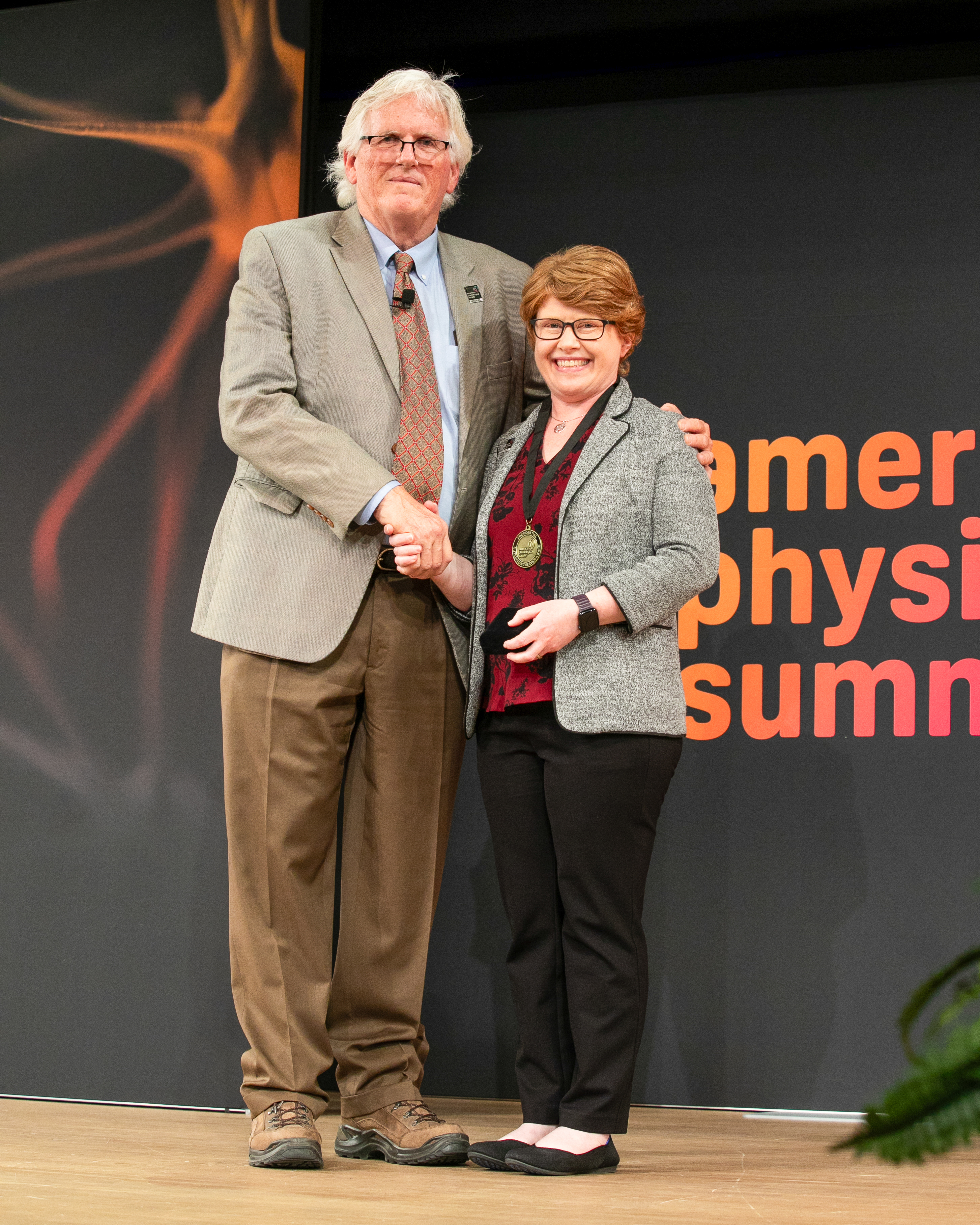 Full uncropped photo of Robert L. Hester, in a blazer, one of the APS Board of Directors and president, shaking hands with UNT Health's Dr. Caroline Rickards, who is wearing her award medal around her neck.