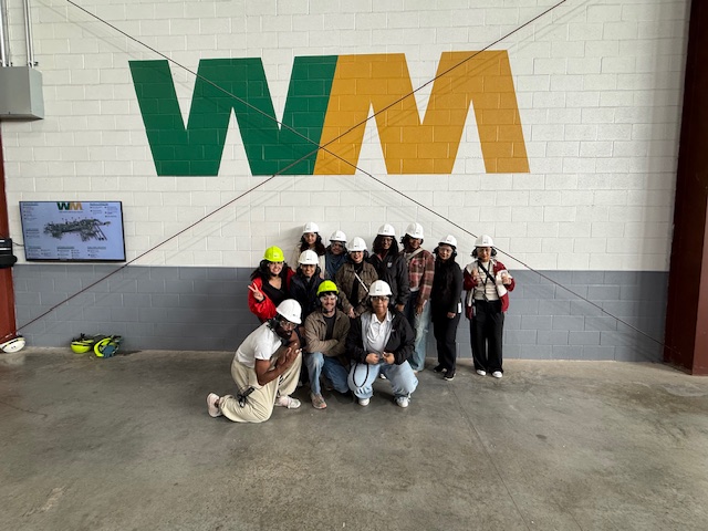 Photo of UNT Health Fort Worth College of Public Health MPH students wearing hard hats pose as a group inside a Waste Management recycling facility in Fort Worth, Texas, with the WM logo painted on the wall behind them.