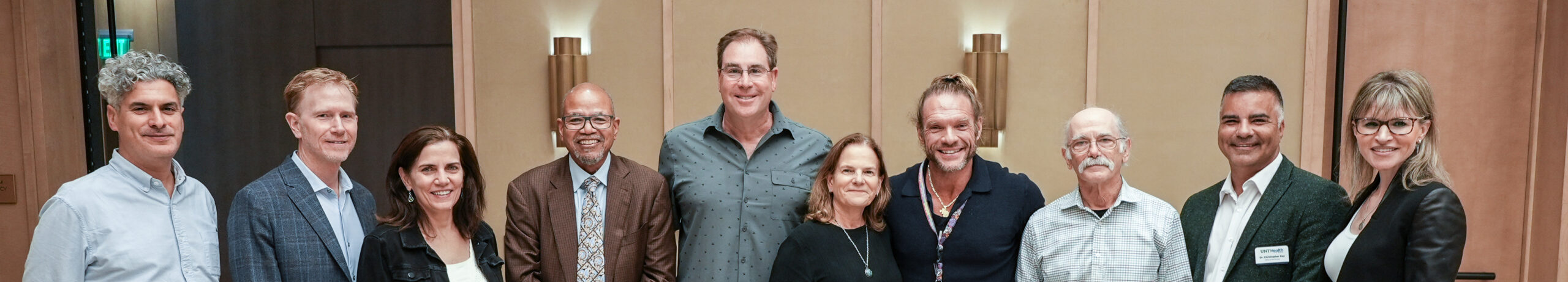 Top horizontal narrow cropped photo banner of a group of nine people standing together in a formal indoor setting, standing together and smiling (fourth from left is UNT Health president, Dr. Calhoun).