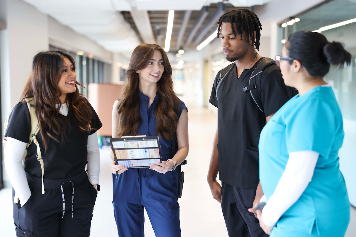 Photo of four UNT Health College of Nursing diverse nursing students in scrubs engage in discussion in a modern hospital hallway, as one student holds a clinical clipboard at UNT Health Fort Worth.