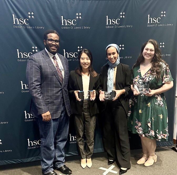 Photo of UNTHSC College of Nursing's Tonychris Nnaka standing next to three women, each holding their own award in front of them in front of a backdrop with a repeated UNTHSC logo pattern printed on it.