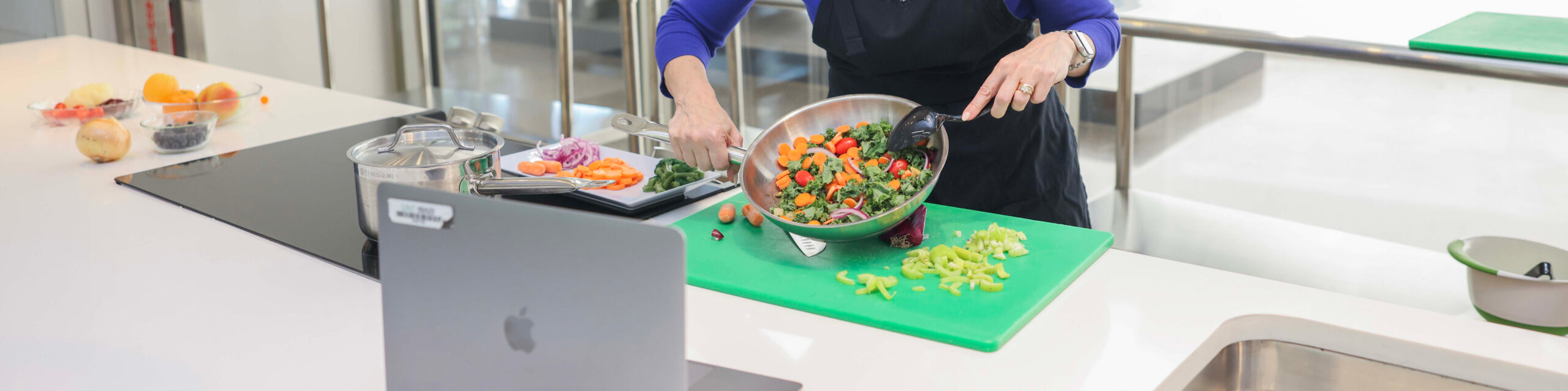 Top cropped horizontal photo banner of a woman wearing an apron at UNT Health is preparing a colorful vegetable dish while engaging in a virtual cooking session via a laptop in a modern kitchen.