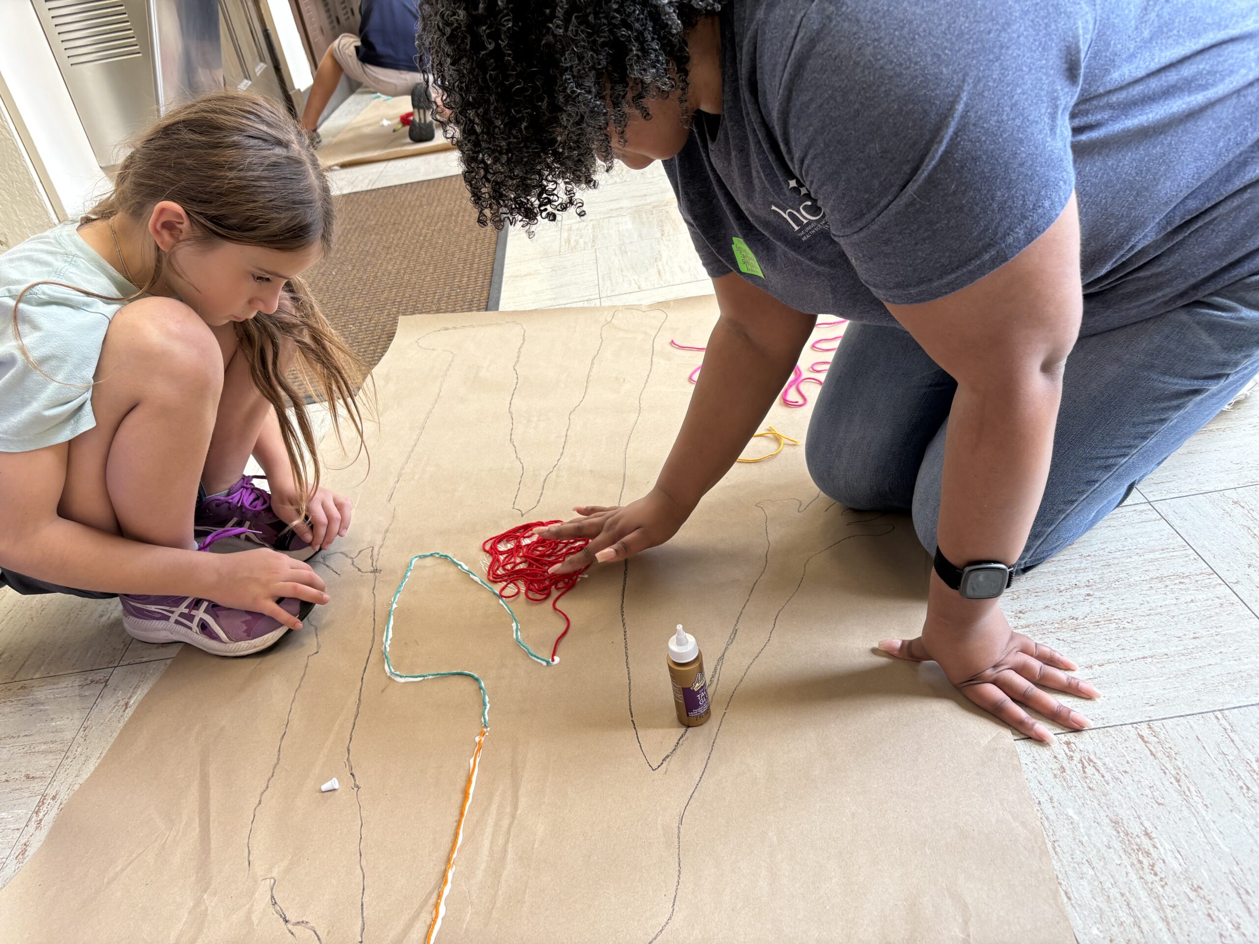 Photo of UNT Health's Kenya Samuels, leading an interactive session at Alice Carlson Elementary School kneeling on the floor together with a element school student with colorful markers, yarn and glue (1 of 2)