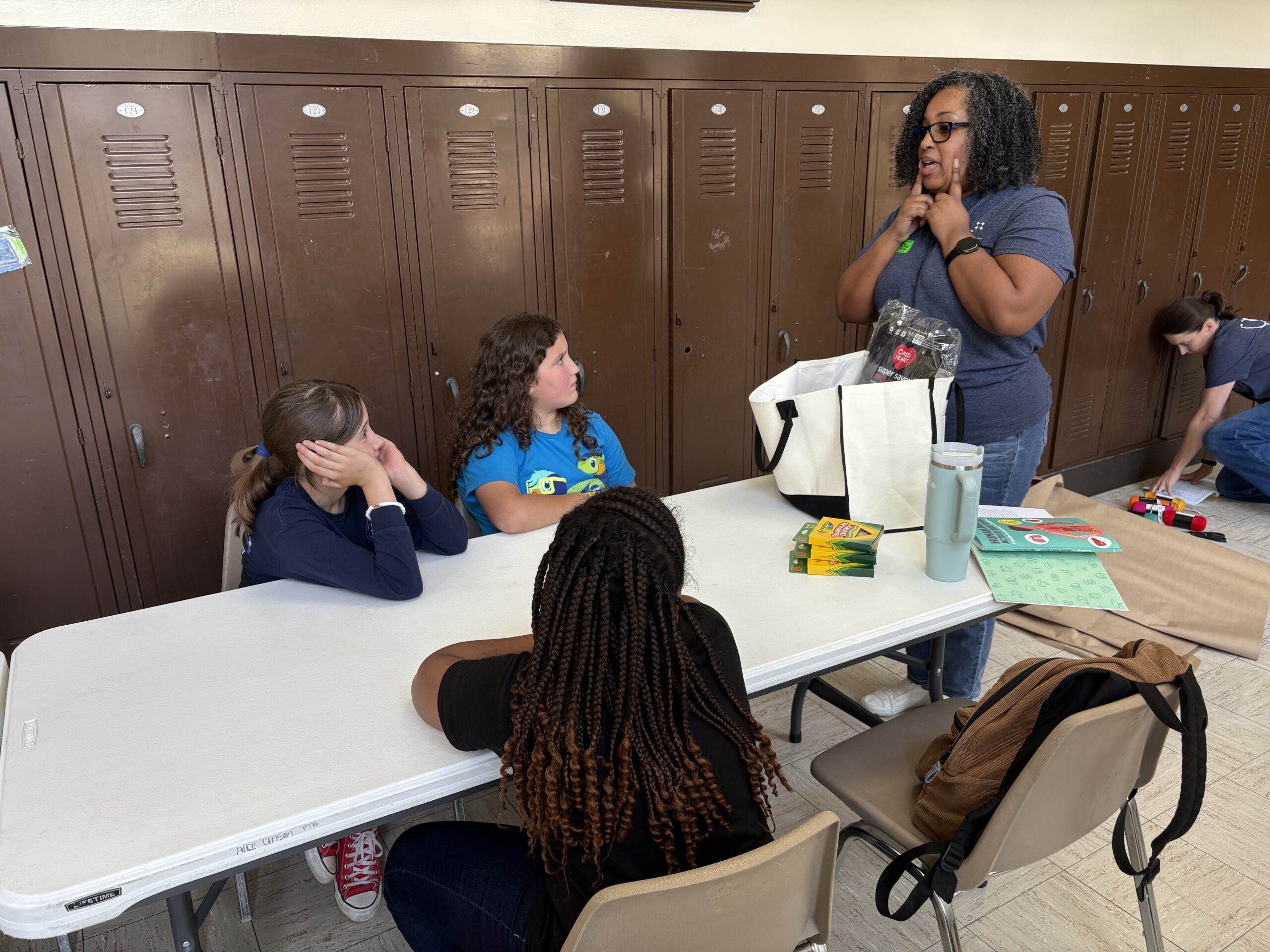 Photo of UNT Health's Kenya Samuels, leading an interactive session at Alice Carlson Elementary School while standing in front of a table with assorted boxes of crayons and a bag. She is talking to three seated students listening (2 of 2)
