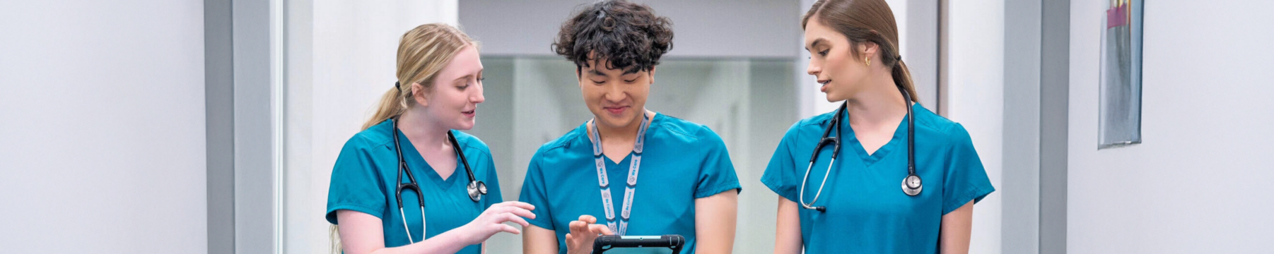 Three nursing students in teal scrubs and stethoscopes walk through a hospital corridor, collaborating over a tablet device at UNT Health Fort Worth. Photo of three nursing students in teal scrubs and stethoscopes walk through a hospital corridor, collaborating over a tablet device at UNT Health Fort Worth.