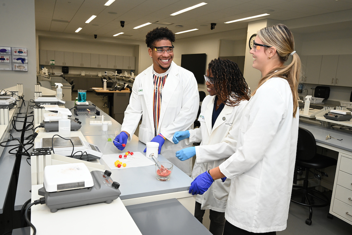 Photo of three UNT Health Fort Worth College of Pharmacy students in white lab coats and blue protective gloves work together in a modern pharmacy skills laboratory, smiling while conducting a hands-on experiment with colorful capsules and compounds.
