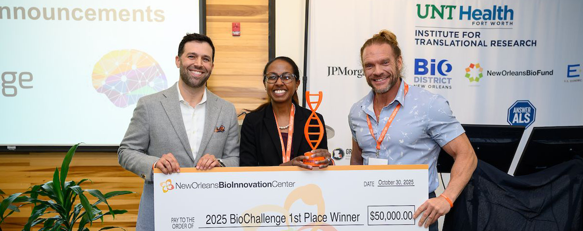 Top horizontal narrow cropped photo banner of three people smiling on stage, holding a large 1st place $50,000 check and the middle woman shows off the check and her trophy at a bio innovation award event. On the left is UNT Health's Dr. Sid O’Bryant, executive director for the Institute for Translational Research.