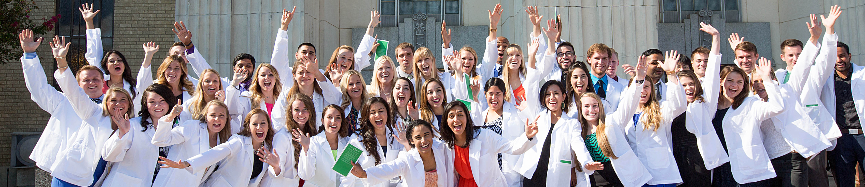 Top narrow horizontal cropped photo banner of a group of smiling UNT Health students wearing their white coats, posing and celebrating outside a campus building.
