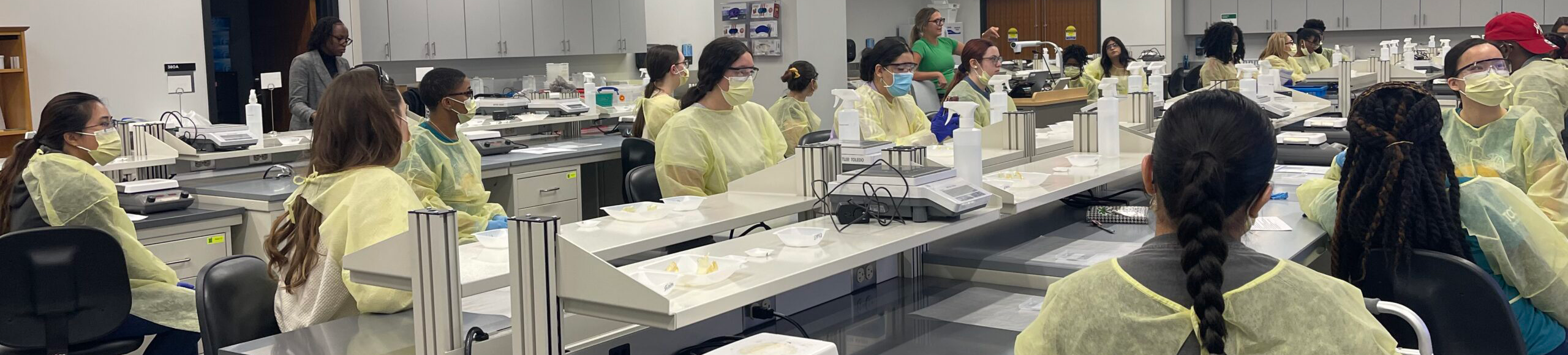 Top horizontal narrow banner of UNT Health's laboratory classroom with students wearing yellow lab gowns and face masks
