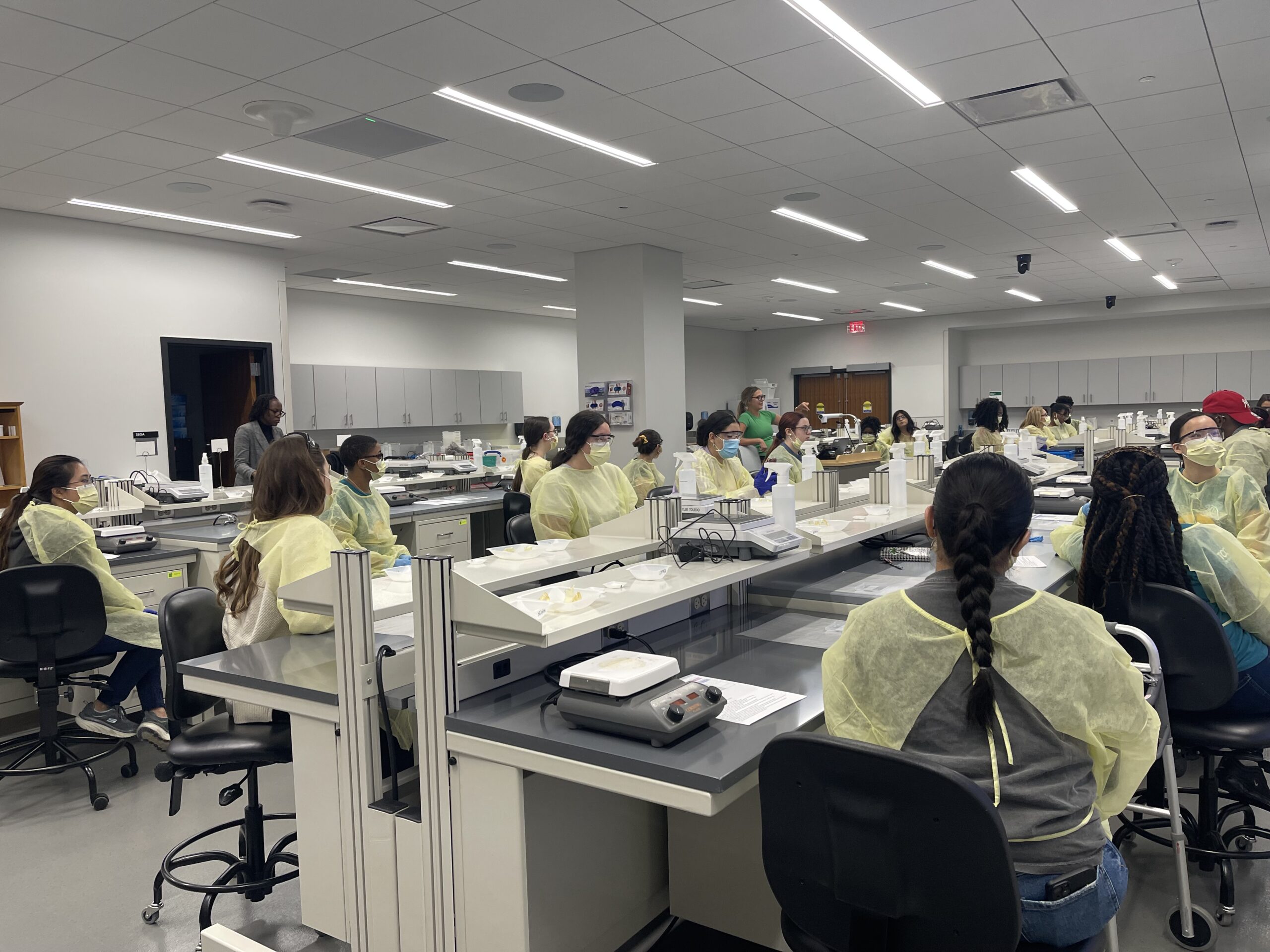 Wide angle reveling students wearing yellow lab gowns and face masks are seated in a brightly lit laboratory classroom. They are listening to instructions from two instructors at the front of the room. Laboratory equipment and materials are arranged on the long white tables with black swivel chairs.